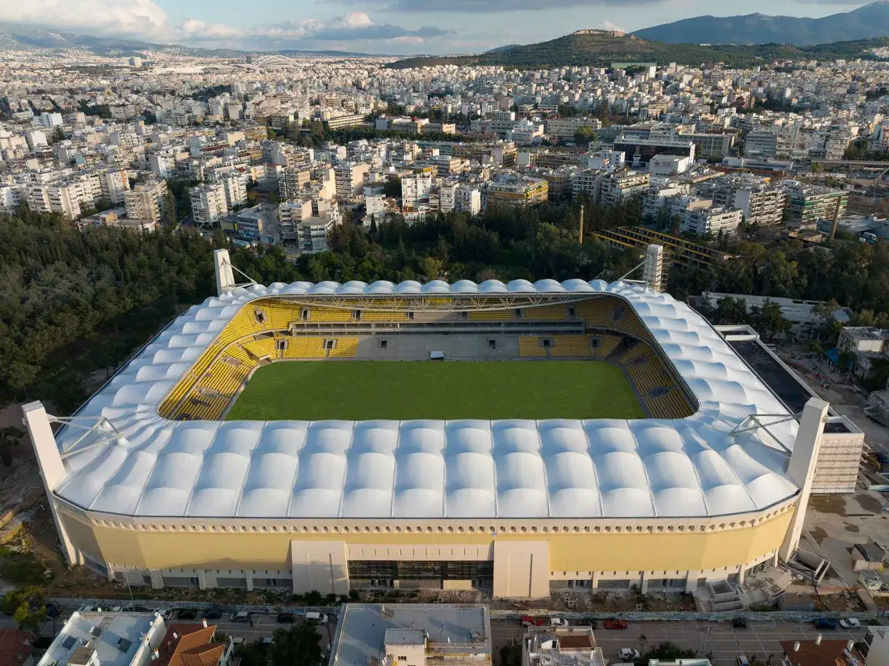Stadion AEK Ateny z żółtymi trybunami i zieloną murawą, otoczony gęstą zabudową miejską i górami w tle.