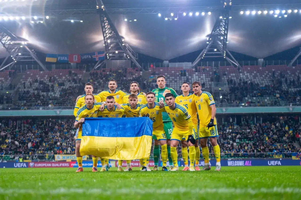 Piłkarze w żółtych strojach z ukraińską flagą, gotowi do meczu. W tle trybuny stadionu i banery UEFA.