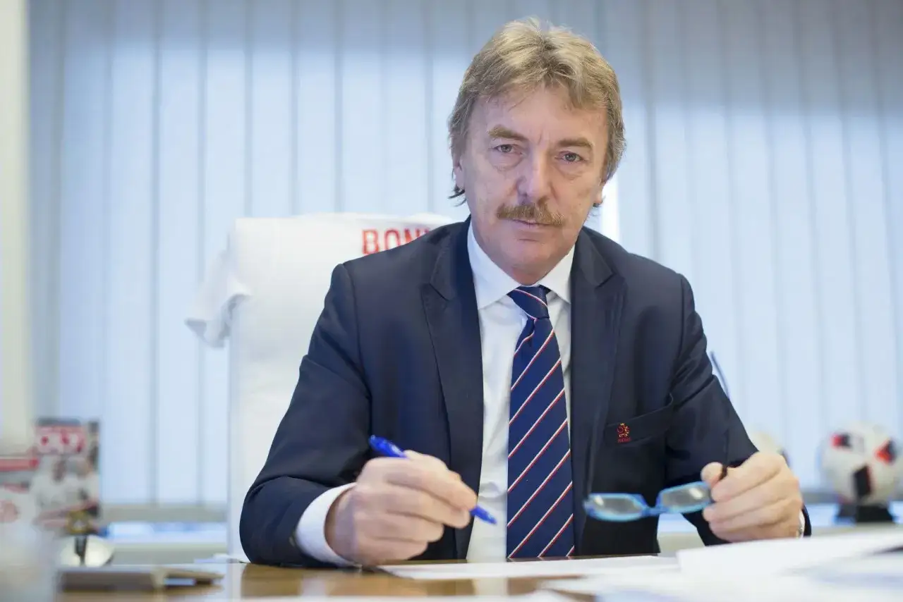 Prezes PZPN w garniturze siedzi przy biurku, trzymając długopis i okulary. W tle biała koszulka z napisem 