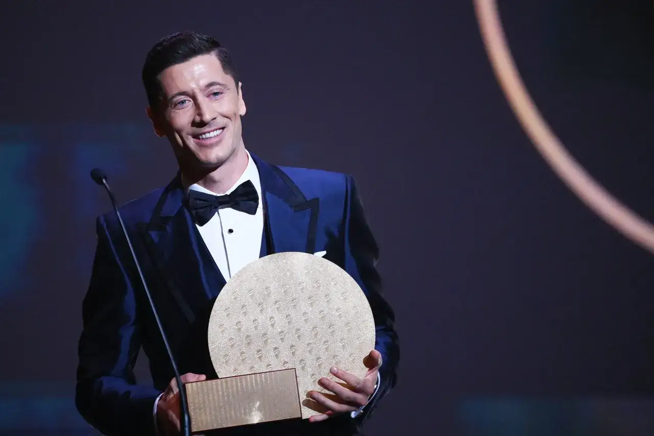 Uśmiechnięty Robert Lewandowski w eleganckim garniturze odbiera nagrodę, być może nawiązującą do jego sukcesów, jak Złota Piłka.